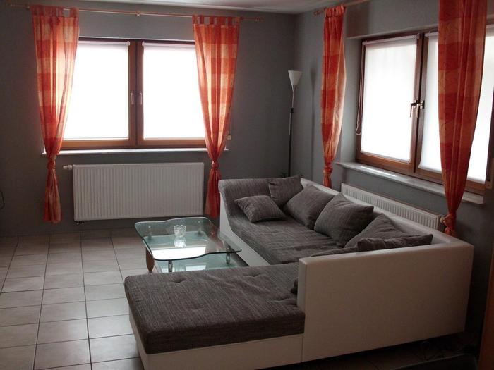 Bild vom Wohnzimmer: Weiße L-Couch mit grauem Polster und orangefarbige Vorhänge, die vier Fensterseiten zusammengebunden sind.