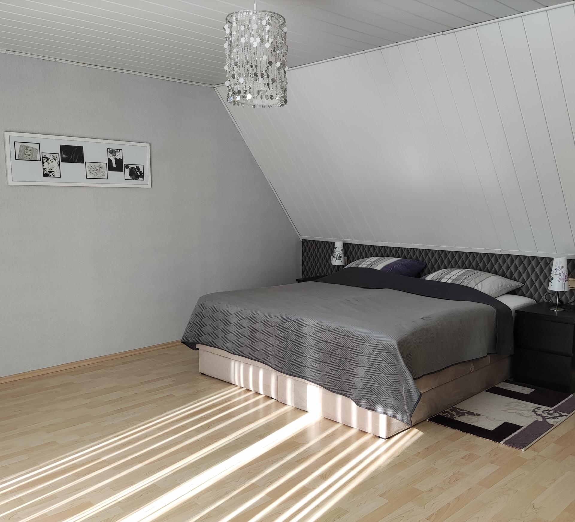 Foto vom Schlafzimmer. Dachsschräge und darunter ein dunkelgraues Doppelbett mit hellgrauer Bettwäsche. Der Boden besteht aus hellem Eichenholz