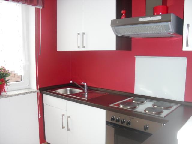 Kleine Weiße Küchenzeile mit einer roten Wand im Hintergrund. Zu sehen ist hier der Spülbecken und ein Cerankochfeld mit einem Backofen unten